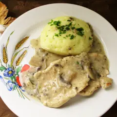 Schab w sosie pieczarkowym z puree ziemniaczanym, ozdobiony natką pietruszki. Danie podane na talerzu z motywem zbóż i kwiatów.