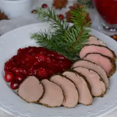 Pokrojona polędwica wieprzowa z sosem żurawinowym i gałązką świerku na białym talerzu.