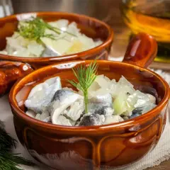 Dwa brązowe naczynia z pysznymi śledziami na słodko, udekorowane gałązkami koperku. Obok widelczyk i butelka z oliwą.