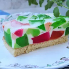 Kolorowe ciasto z galaretkami w kostkę, z zielonymi i różowymi kawałkami zatopionymi w białej masie, na spodzie biszkoptowym.