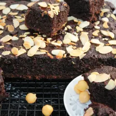 Pyszne brownie z ciecierzycy, posypane płatkami migdałów, wygląda apetycznie na kratce. Kilka kawałków podano na talerzyku z dodatkiem orzechów.