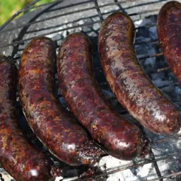 Pięć ciemnych, rumianych kiełbasek na ruszcie grilla. Widać dymek i żar, co sugeruje, jak powstaje kaszanka.