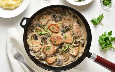 Poledwiczki wieprzowe w sosie pieczarkowym, podane w czarnej patelni, ozdobione rozmarynem i natką pietruszki. Obok puree ziemniaczane.