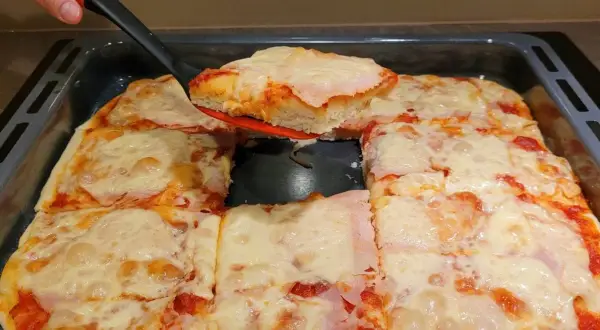 Pyszna, domowa pizza z blachy, z ciągnącym się serem i szynką, właśnie kroimy pierwszy kawałek.