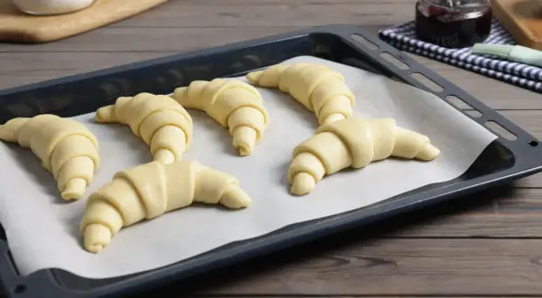 Sześć surowych rogalików z ciasta francuskiego czeka na pieczenie. Obok słoiczek z dżemem.