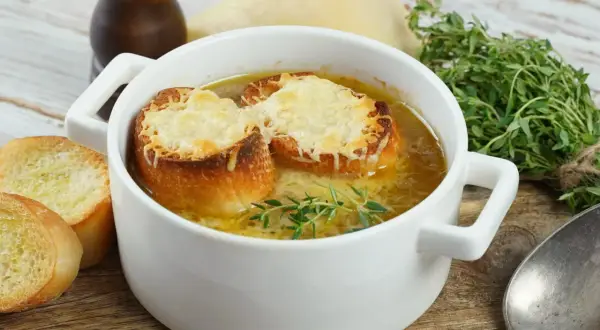 Jak zrobić zupę cebulową? Aromatyczna zupa cebulowa z grzankami i serem, podana w białej miseczce, z gałązką tymianku.