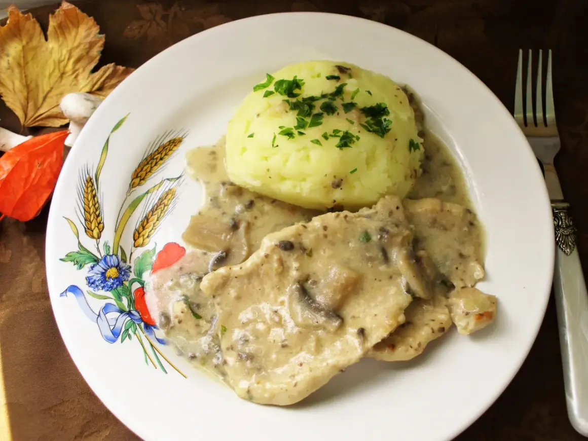 Schab w sosie pieczarkowym z puree ziemniaczanym, ozdobiony natką pietruszki. Danie podane na talerzu z motywem zbóż i kwiatów.