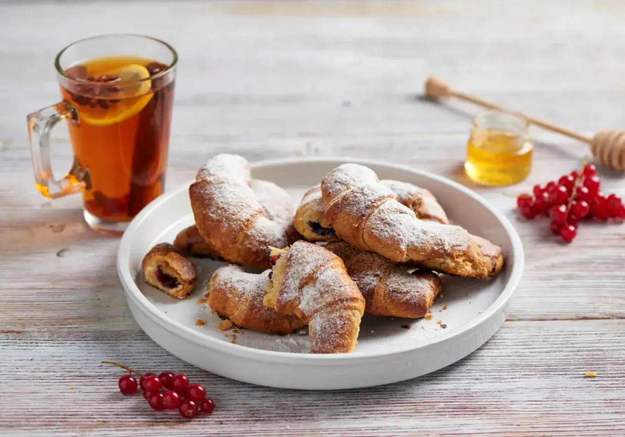 Pyszne rogaliki z ciasta francuskiego z dżemem, posypane cukrem pudrem, obok kubka herbaty i czerwonych porzeczek.