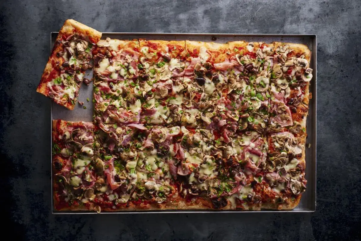 Pyszna domowa pizza z blachy, obficie posypana szynką, pieczarkami i oliwkami, z ciągnącym się serem.