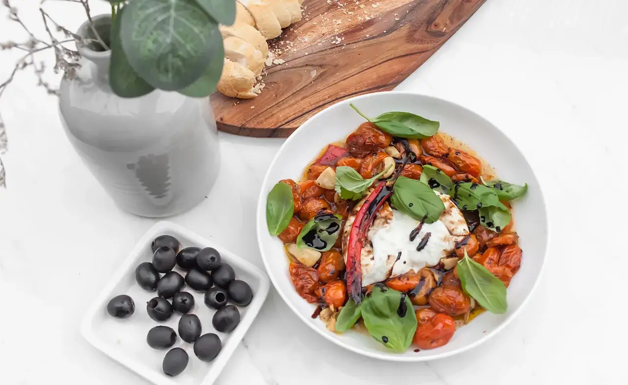 Pyszna burrata z piekarnika z pieczonymi pomidorkami, czosnkiem i bazylią, podana z oliwkami i pieczywem.