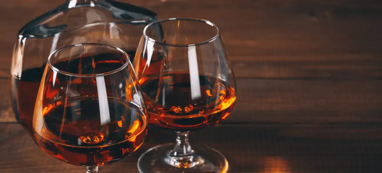 Dwa kieliszki z bursztynowym płynem i karafka na drewnianym tle. Czy wiesz, czym się różni brandy od koniaku?