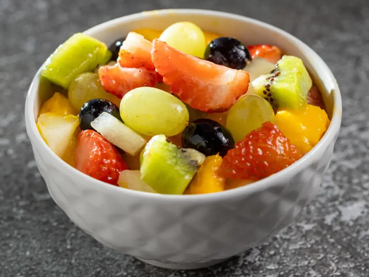 Kolorowa sałatka z owocami: truskawki, kiwi, winogrona, mango i czarne jagody w białej miseczce.